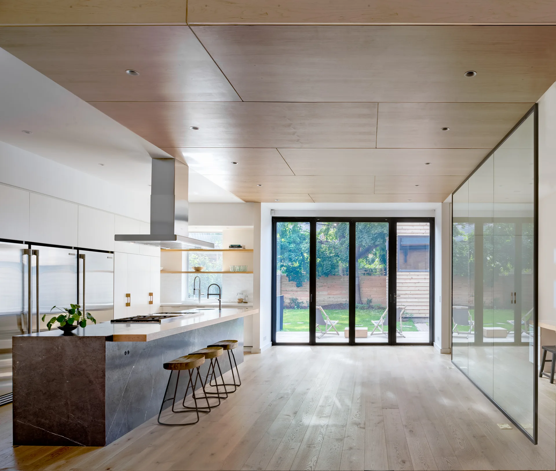 A modern kitchen featuring a dark stone island with wood stools, white cabinetry, a plywood paneled ceiling, and large glass patio doors.