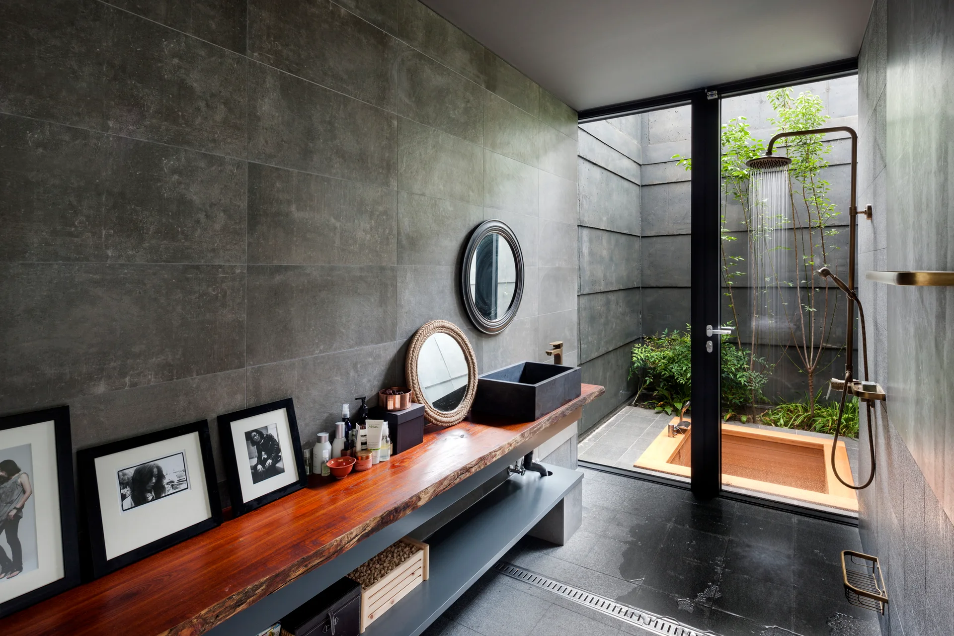 A modern bathroom with dark stone tiles, a long live-edge wood vanity with a black vessel sink, and a glass door opening to an outdoor tub.
