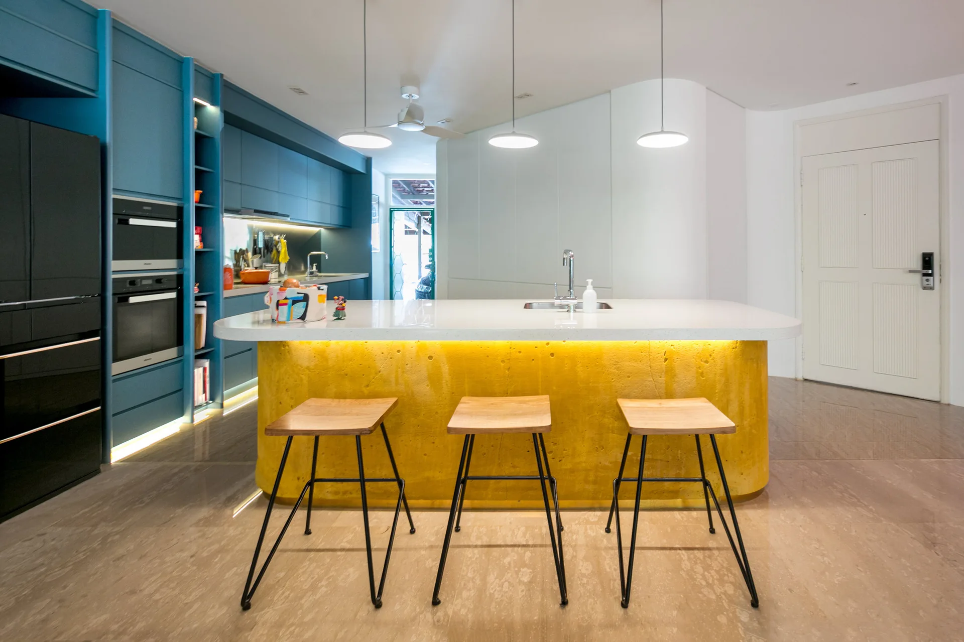 Matte blue cabinetry pairs with a bright yellow textured island base and three wooden bar stools in a contemporary kitchen.