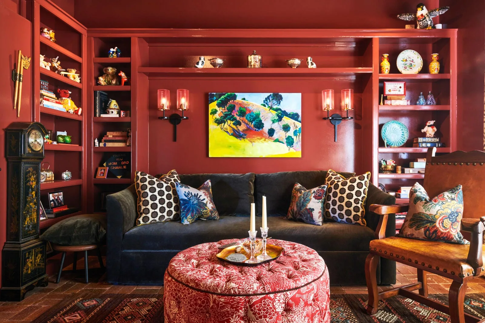 A living room featuring deep red built-in bookshelves, a dark velvet sofa, a patterned red ottoman, and a colorful landscape painting.