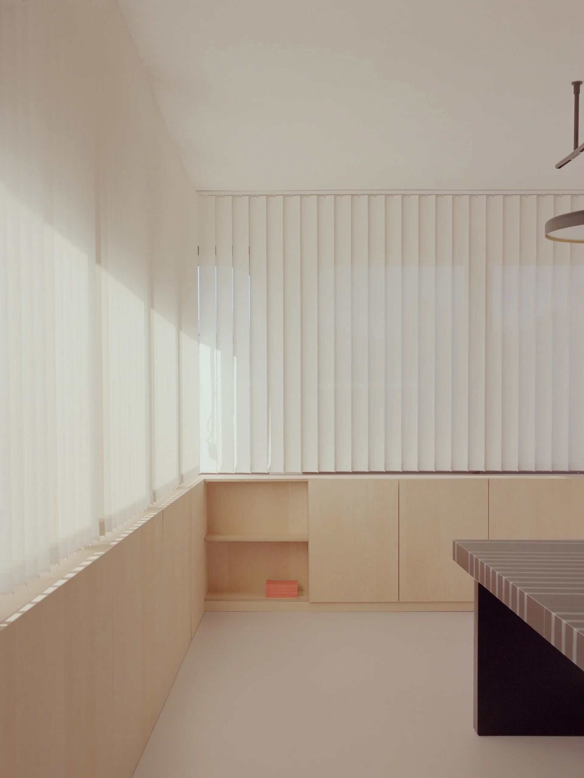 Low-profile light wood cabinets line the walls of a minimalist room beneath windows covered by tall white vertical blinds.