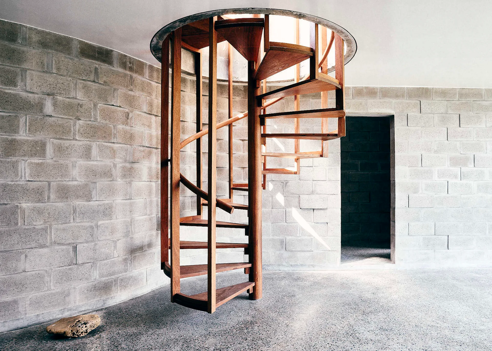 A wooden spiral staircase rises toward a circular ceiling opening in a room with grey concrete block walls and polished floors.