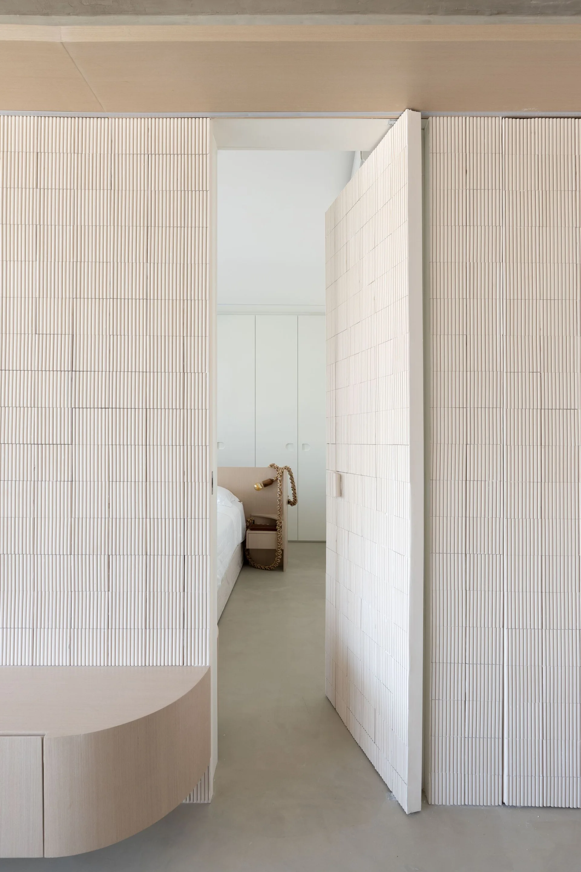 An open doorway made of vertical ribbed paneling leads into a minimalist bedroom featuring a light wood nightstand and built-in white closets.