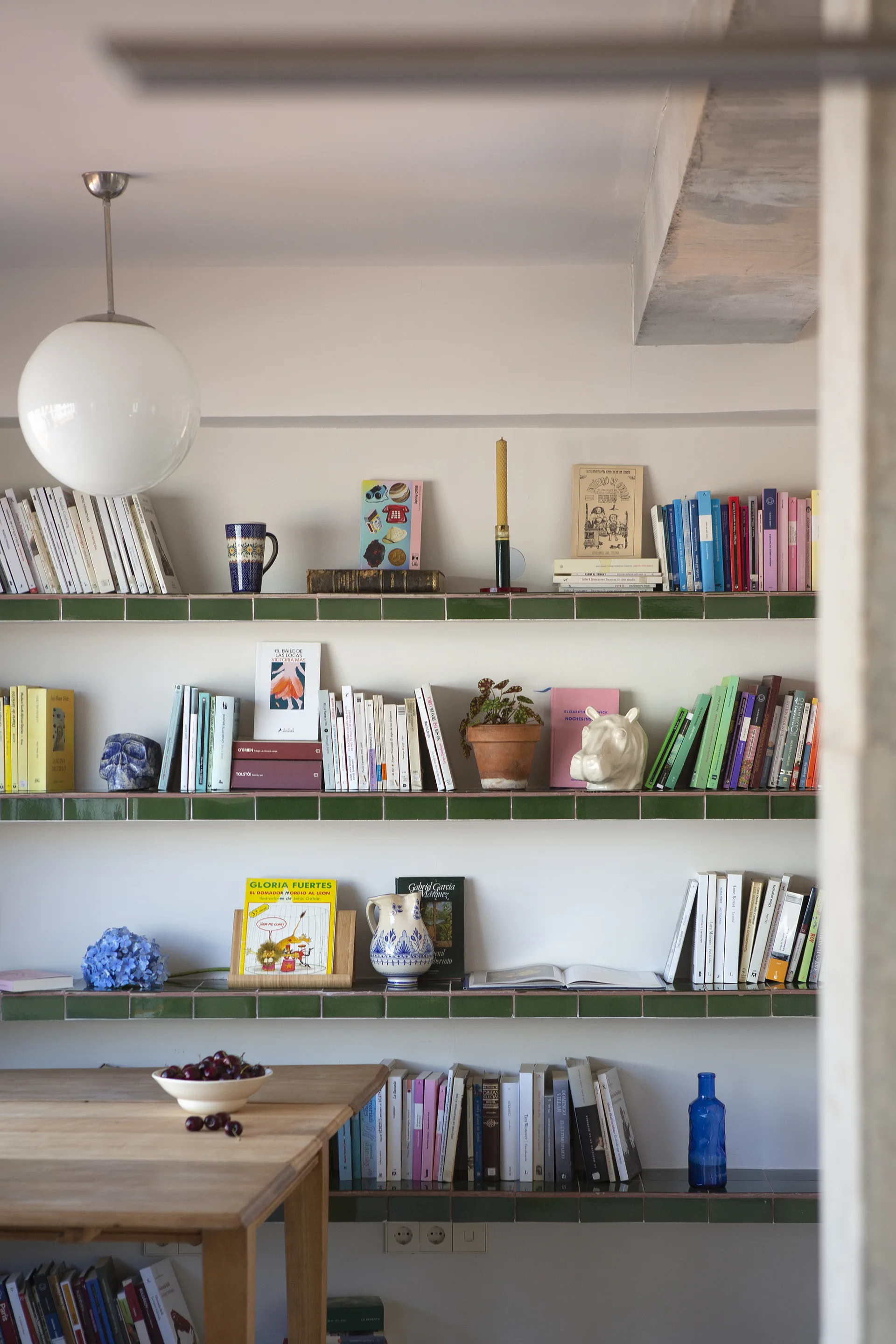 A multi-tiered white bookshelf featuring glossy green tiled edges is styled with books, plants, and art objects above a wooden table.