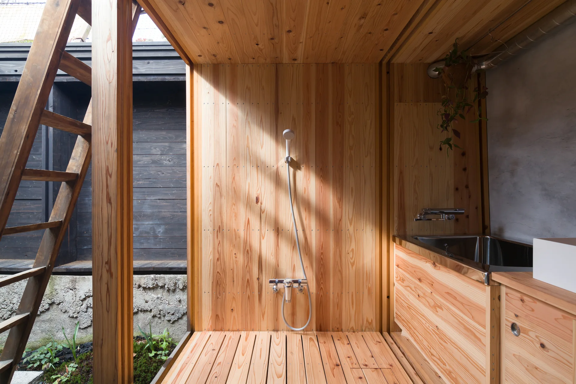 A minimalist wood-paneled shower stall features a hand shower, slatted wood floor, and an adjacent stainless steel sink with a concrete wall.