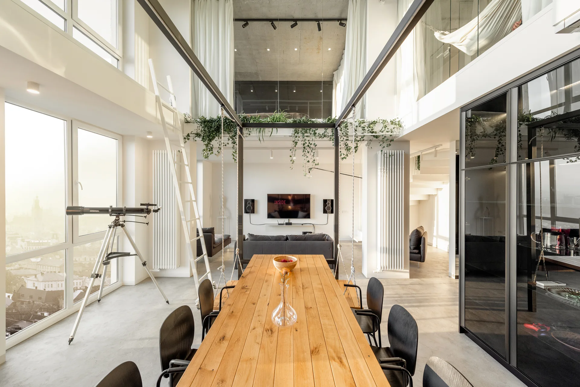A modern loft interior features a long wooden dining table, black chairs, a mezzanine level with trailing plants, and a telescope near tall windows.