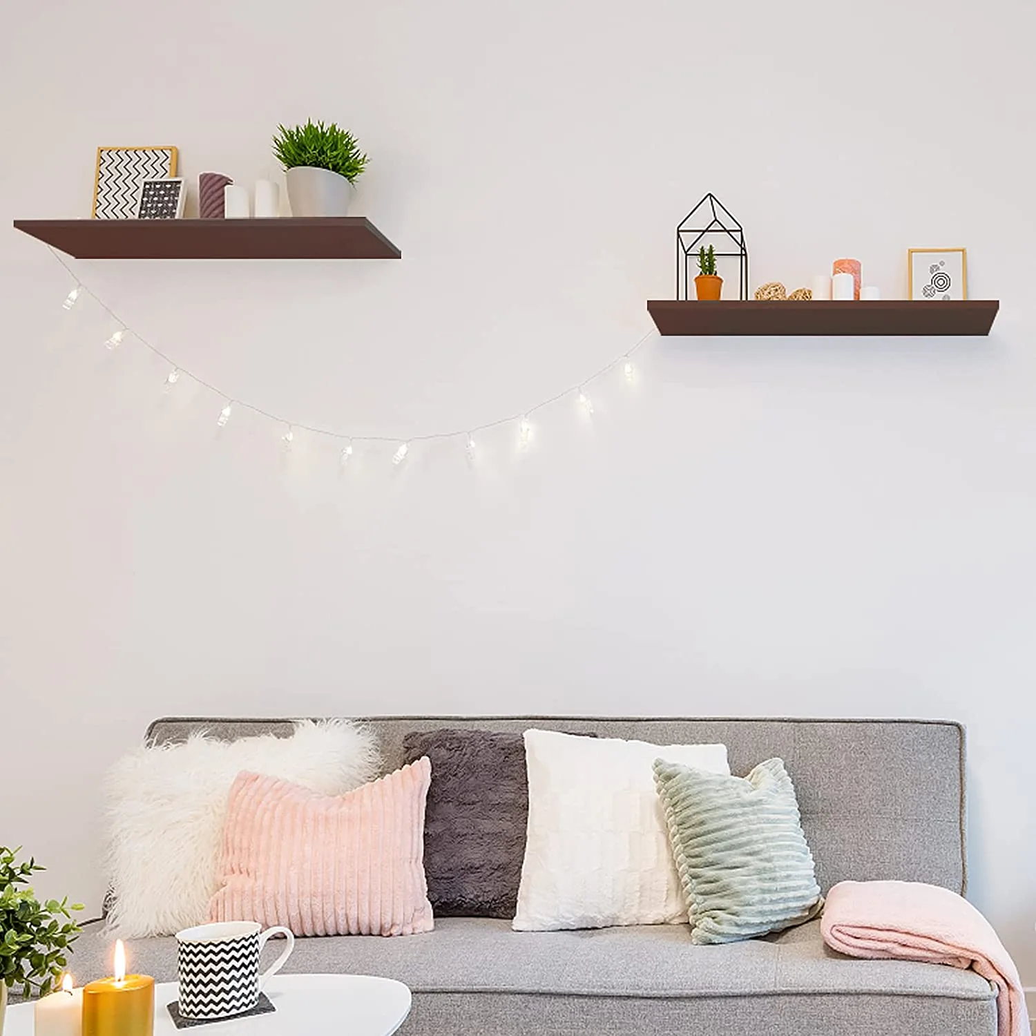 A grey sofa with pink and sage green cushions sits beneath two staggered dark brown floating shelves and draped string lights.