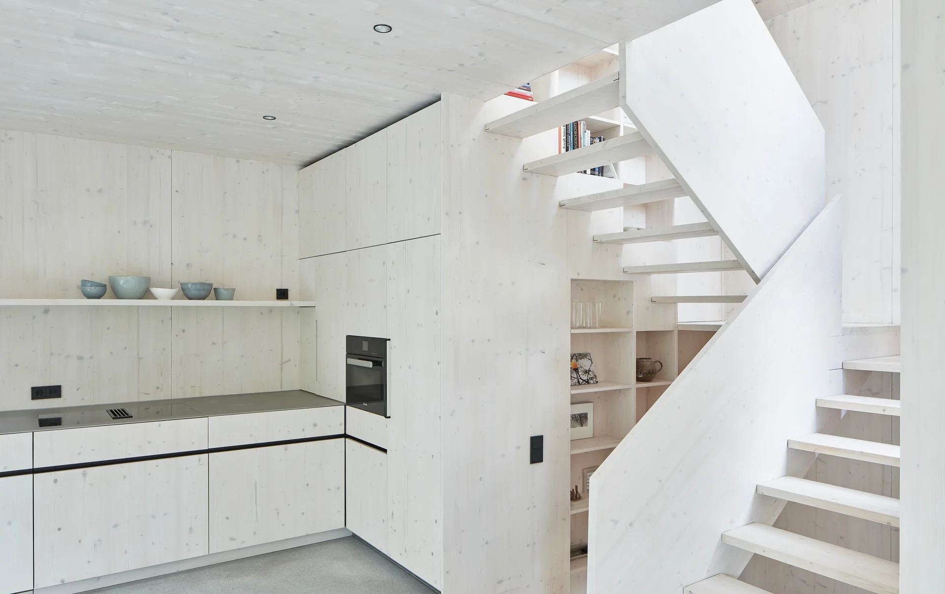 A minimalist kitchen features whitewashed wood cabinetry and walls next to a matching light wood staircase with built-in shelving.