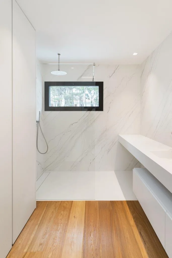 A modern bathroom featuring a walk-in shower with marble tiled walls, a black-framed window, a white floating vanity, and hardwood flooring.