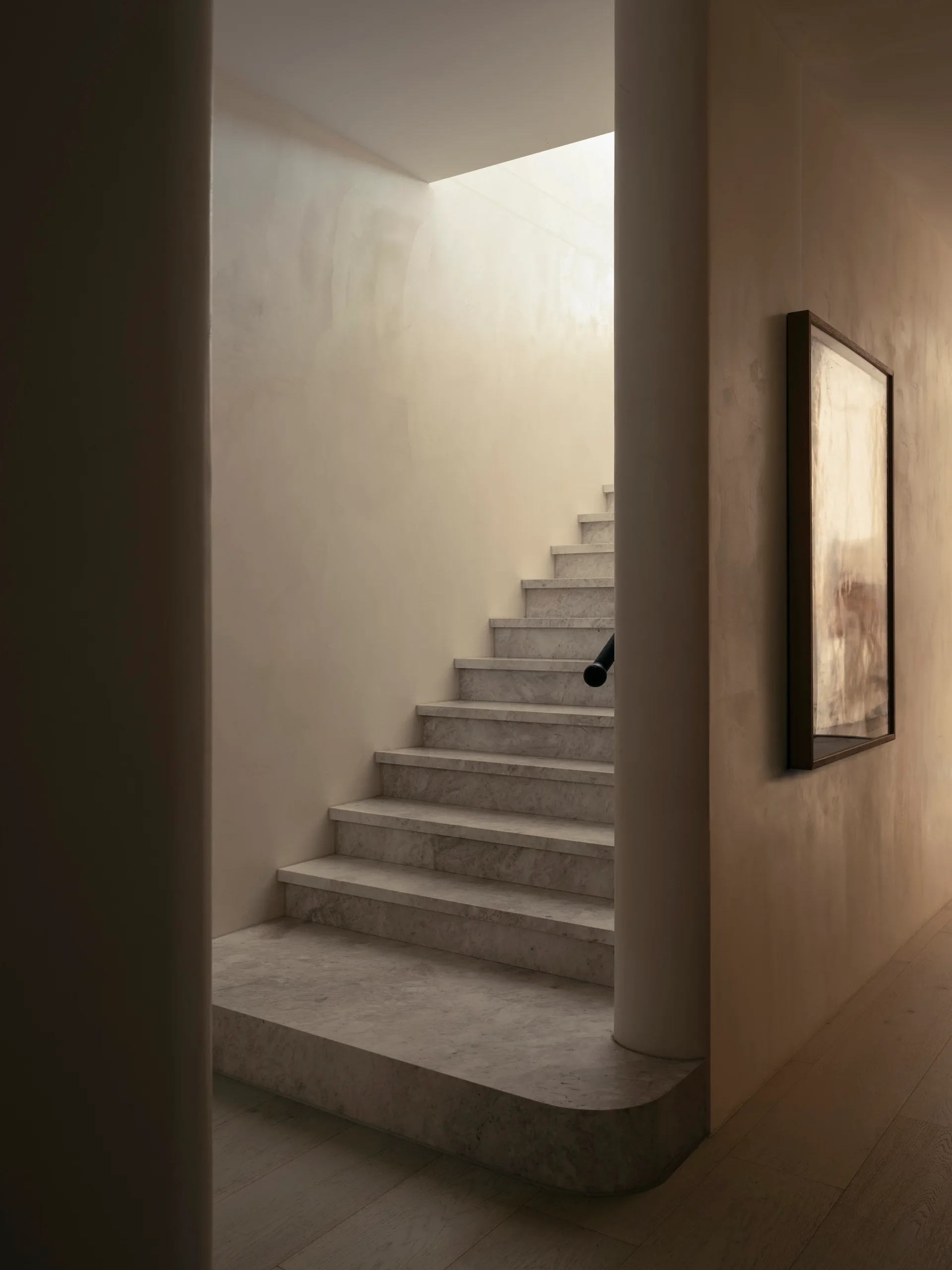 A minimalist grey stone staircase is framed by smooth beige plaster walls under a bright natural skylight, with a framed artwork on the right hallway wall.
