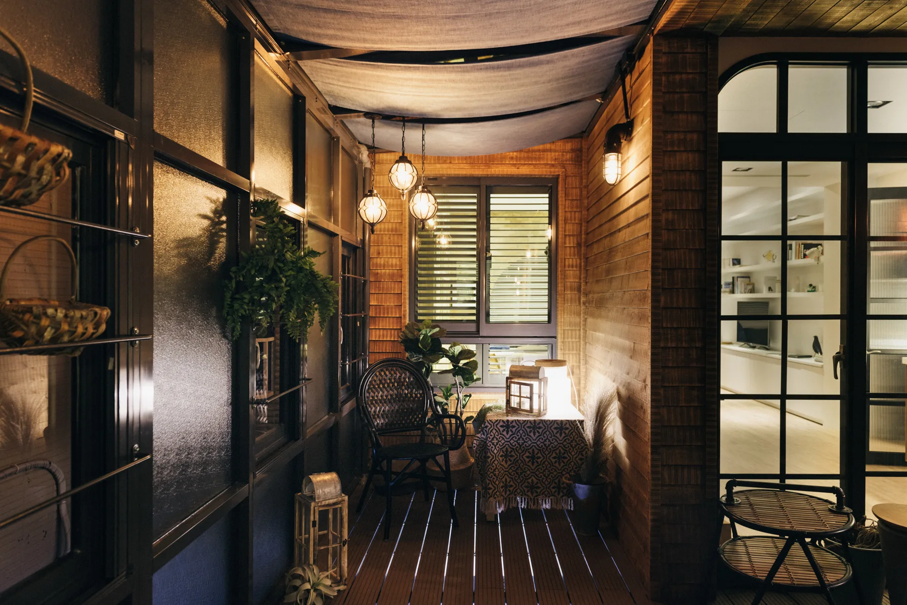 A narrow enclosed balcony featuring warm wood paneled walls, a draped fabric ceiling canopy, a wicker chair, and glowing lanterns.