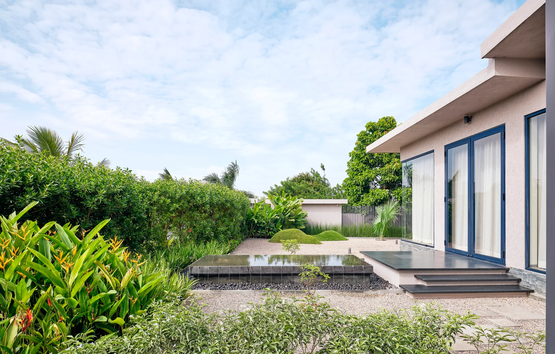 A modern house facade with glass doors faces a gravel garden featuring a low rectangular stone water fountain and lush green hedges under a blue sky.