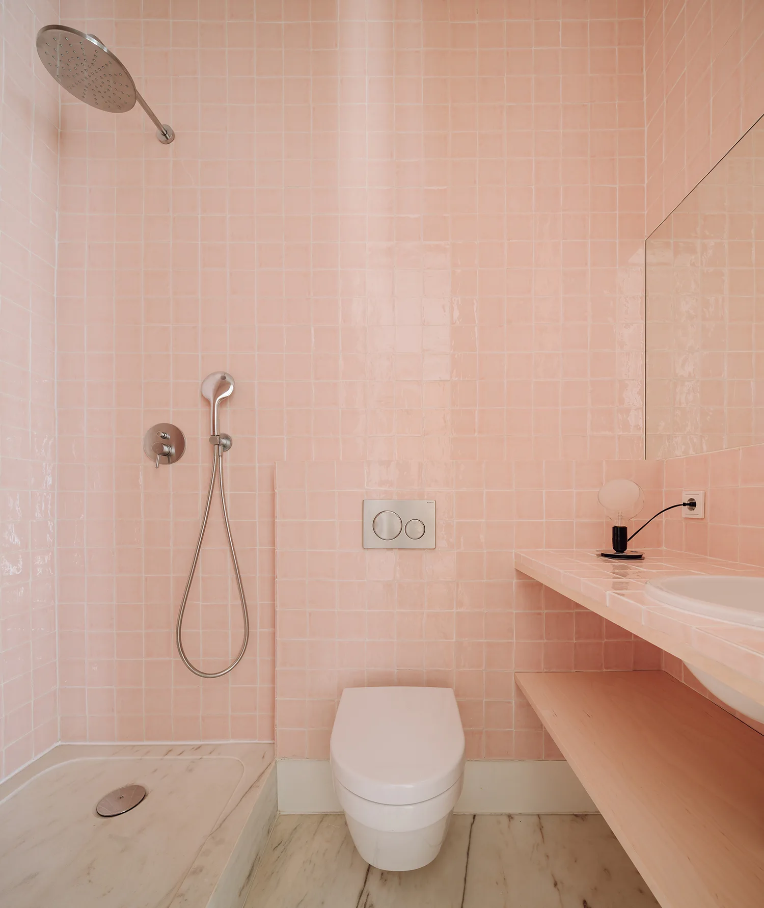 A modern bathroom with glossy pink square wall tiles, a wall-mounted toilet, marble flooring, and an open wooden vanity shelf with an integrated sink.