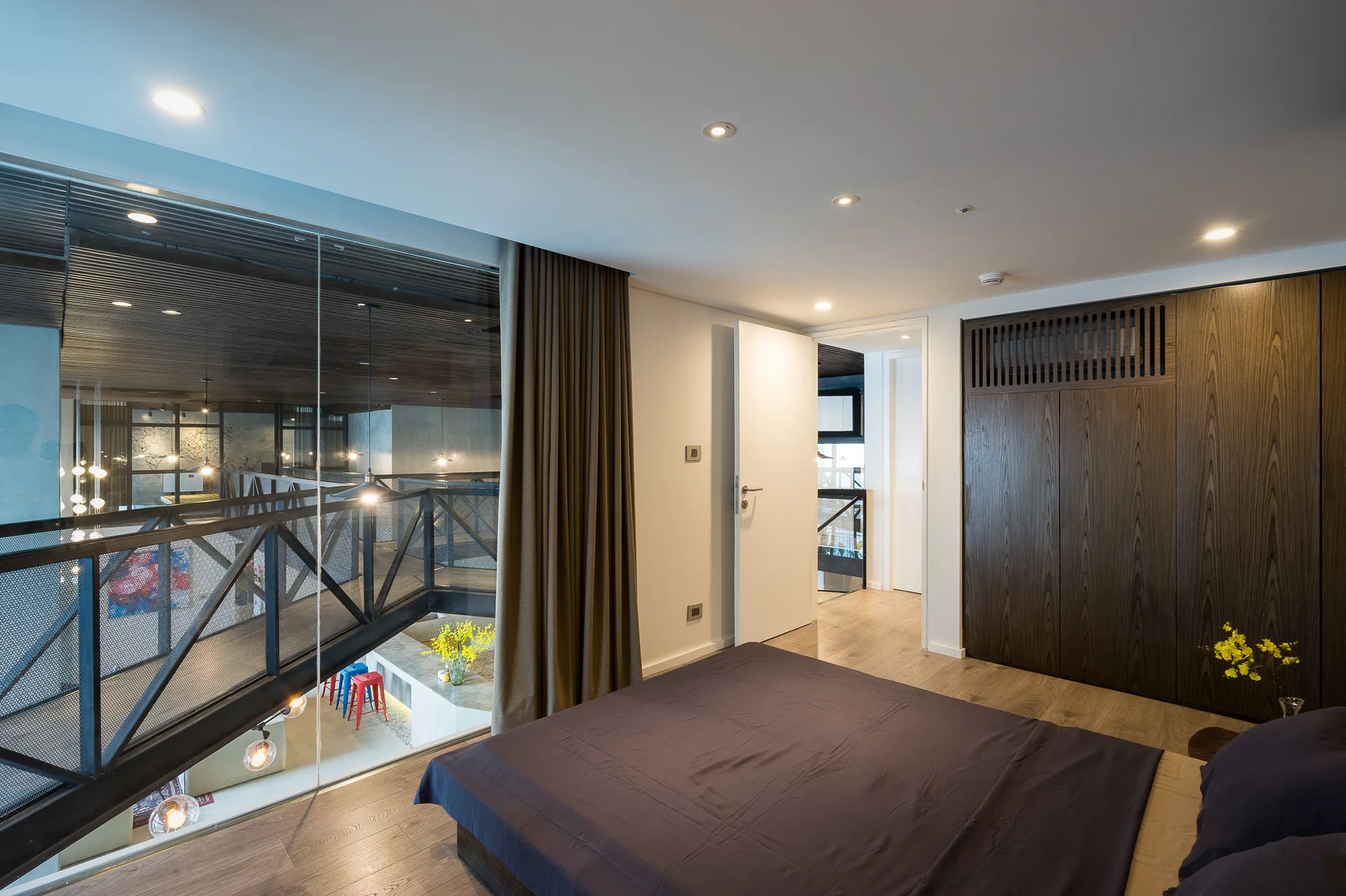 A mezzanine bedroom features a large glass wall overlooking a lower level, a bed with dark bedding, and a built-in dark wood closet.