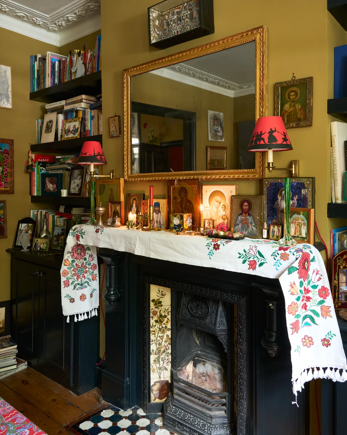 A black fireplace mantel draped in a floral runner is decorated with religious icons and flanked by bookshelves against green walls.