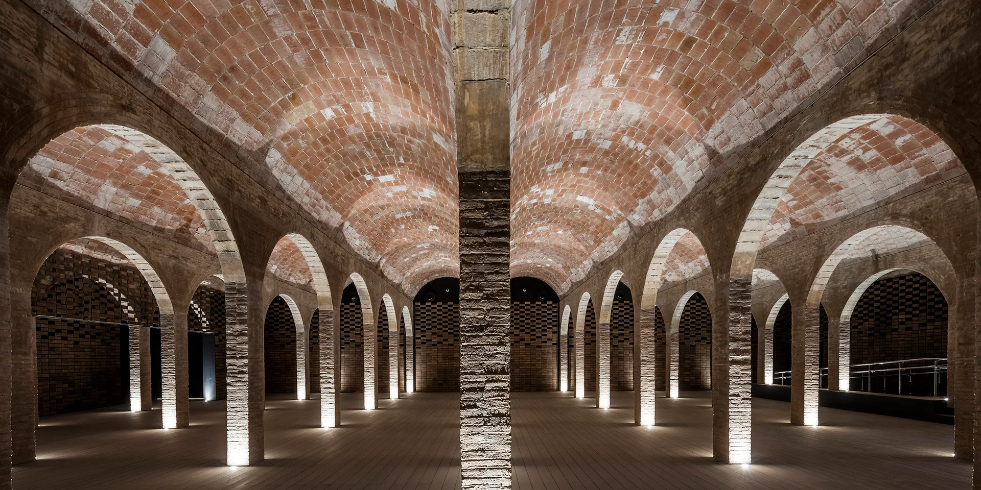 A spacious interior hall with repeating red brick archways, vaulted ceilings, and wooden floors illuminated by floor uplights.