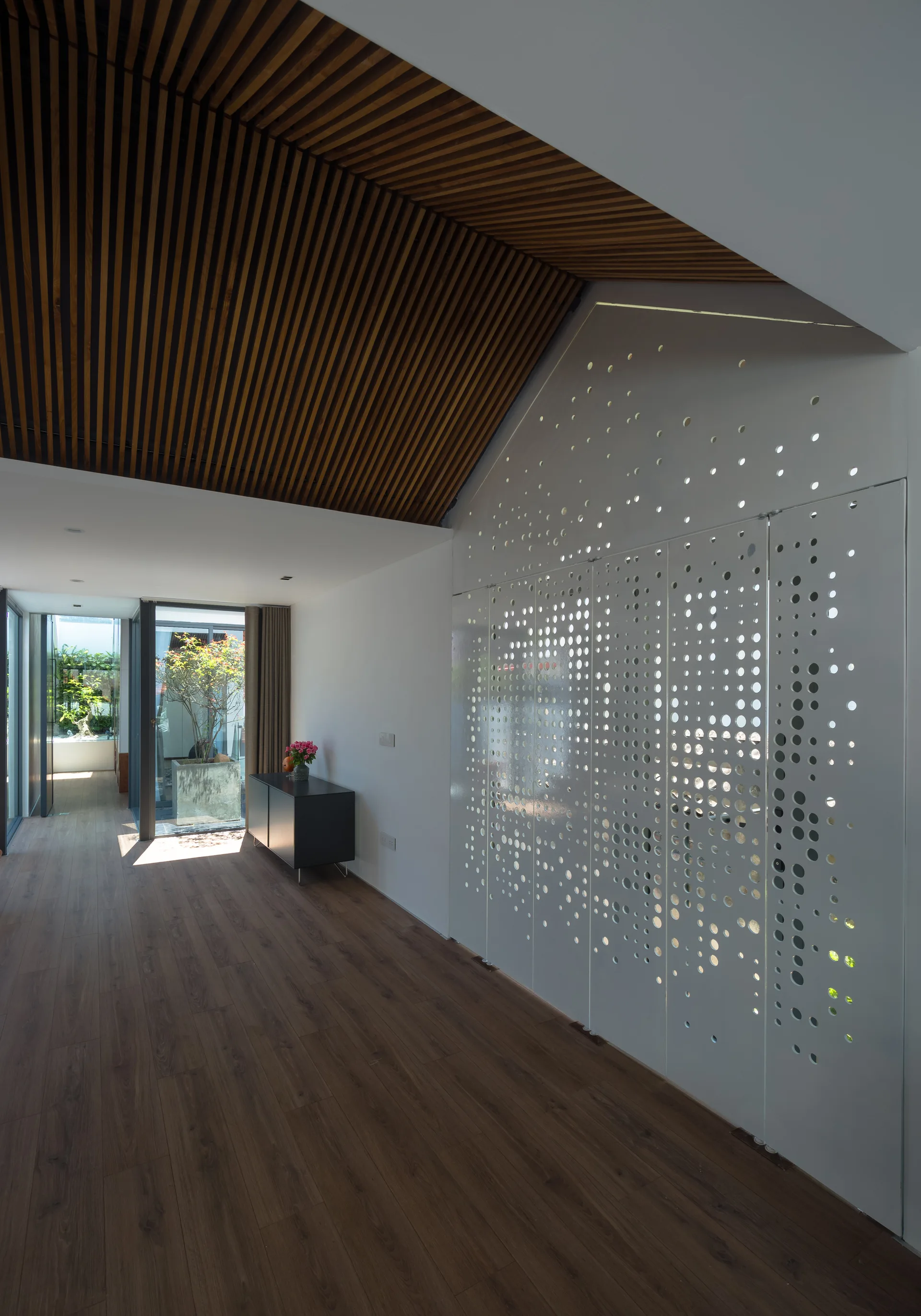 A modern hallway featuring a sloped wood-slatted ceiling, a white perforated metal partition wall, and dark wood flooring leading to a sunlit courtyard.