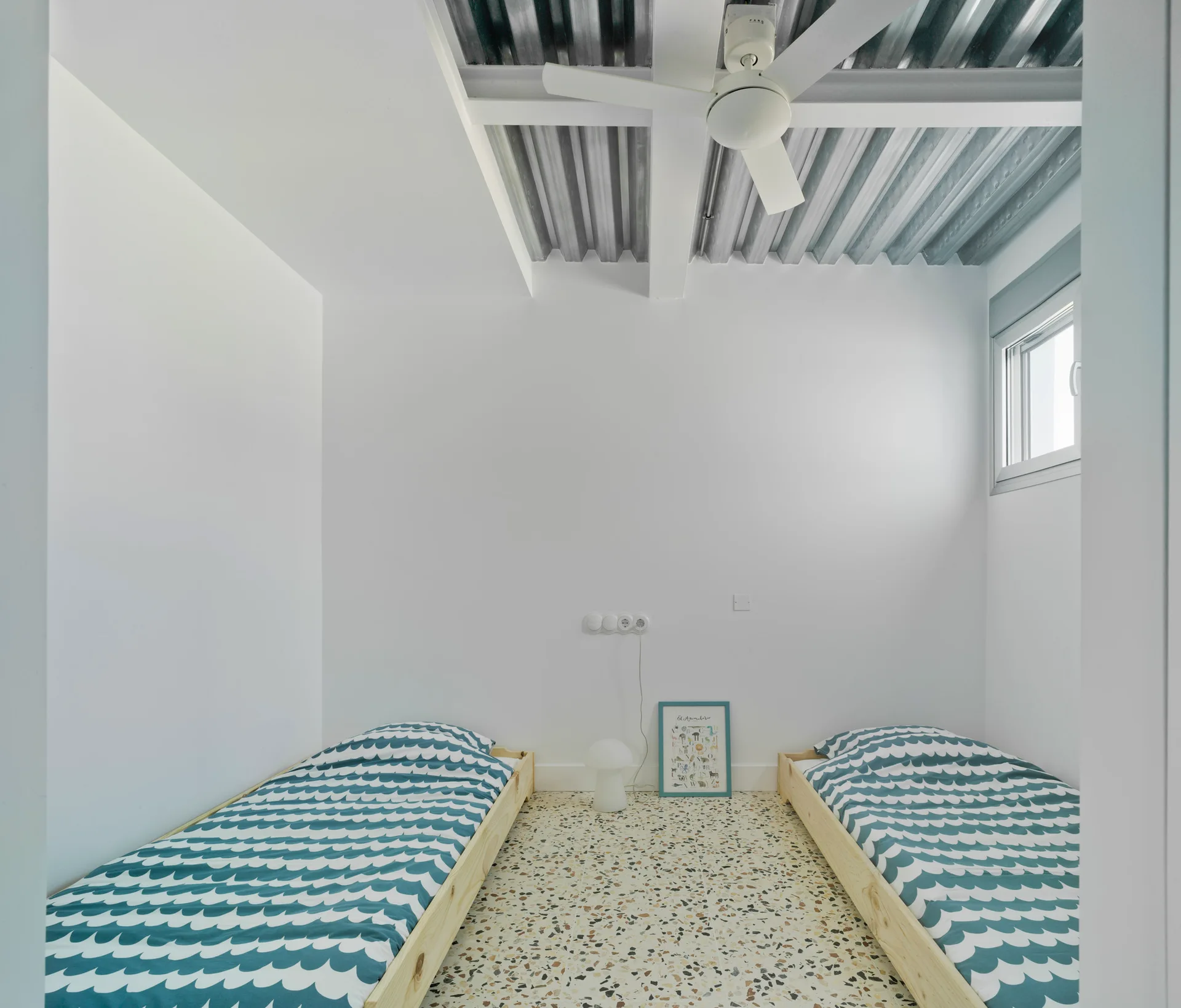 Two low pine twin beds with blue patterned sheets sit on a speckled terrazzo floor under an industrial corrugated metal ceiling.
