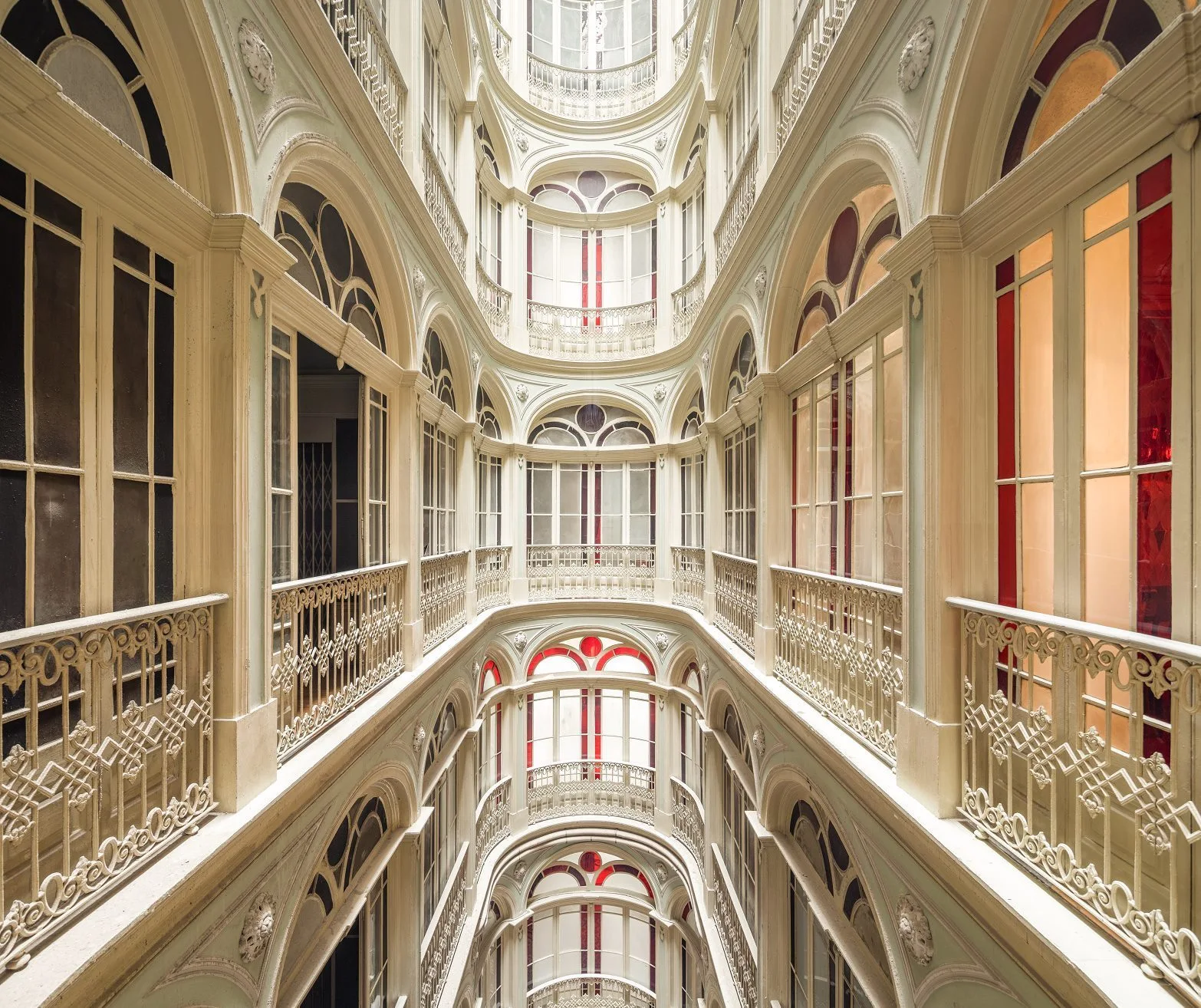 An interior multi-level atrium featuring cream plaster arches, decorative metal railings, and small red stained glass window panes.