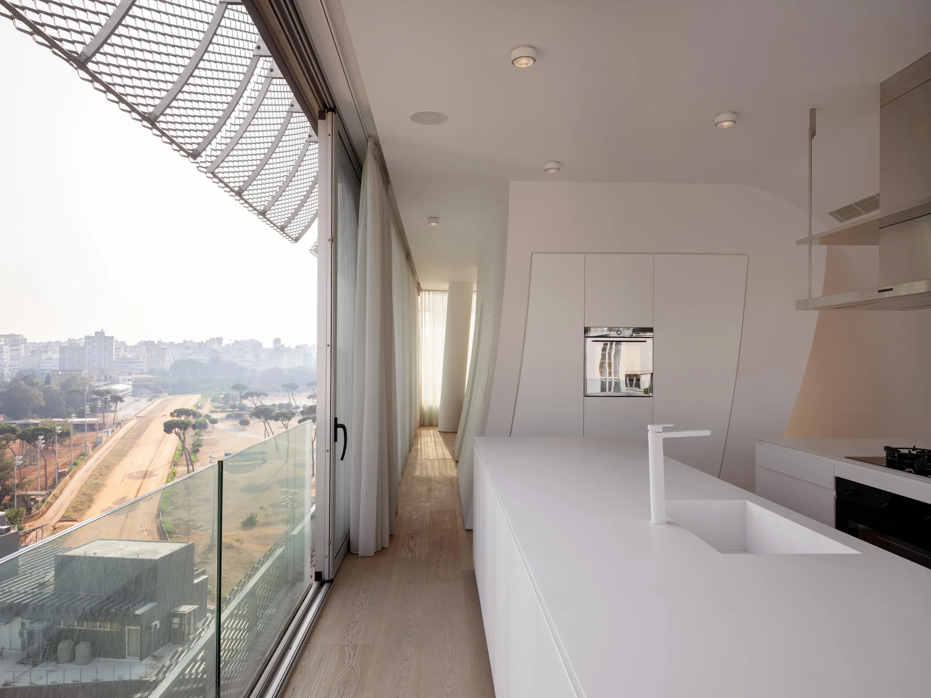A minimalist white kitchen features a long central island with a matching faucet, next to sliding glass doors that open to an outdoor park view.