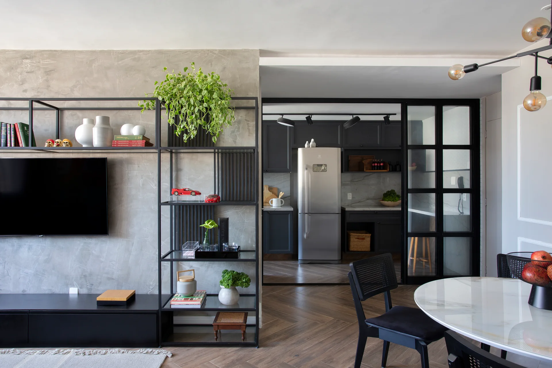 A living room with a textured grey concrete wall, black steel shelving, and a glass-paned sliding partition that reveals a dark kitchen.