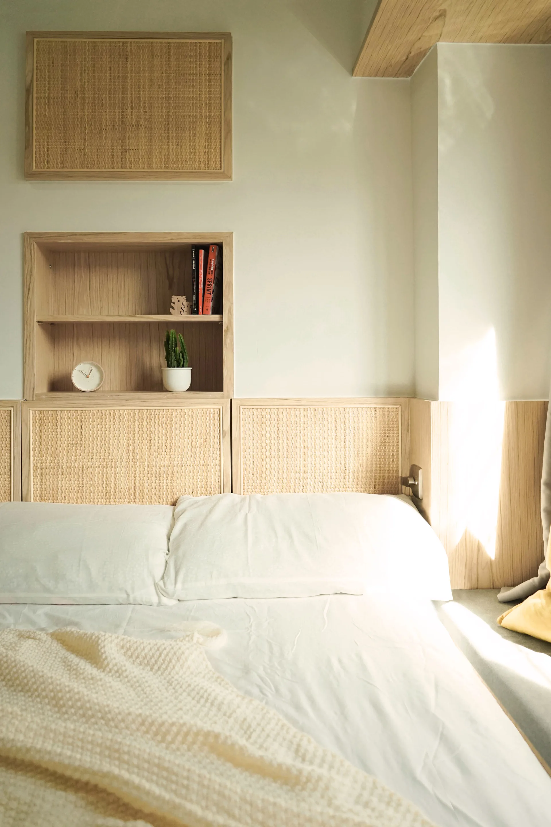 A bedroom features white bedding and a custom light wood headboard with woven rattan panels and a recessed display niche holding books and a small plant.