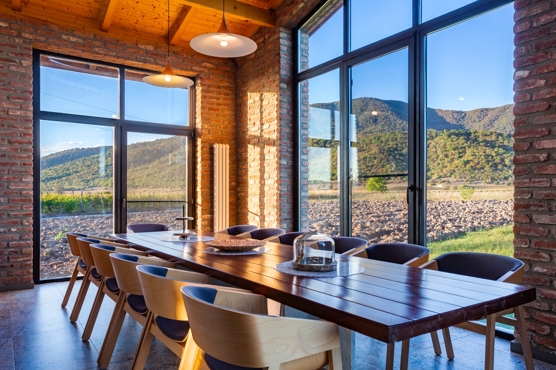 A long wood dining table surrounded by curved chairs sits in a room with exposed brick walls, wood-beamed ceilings, and large windows showing mountains.