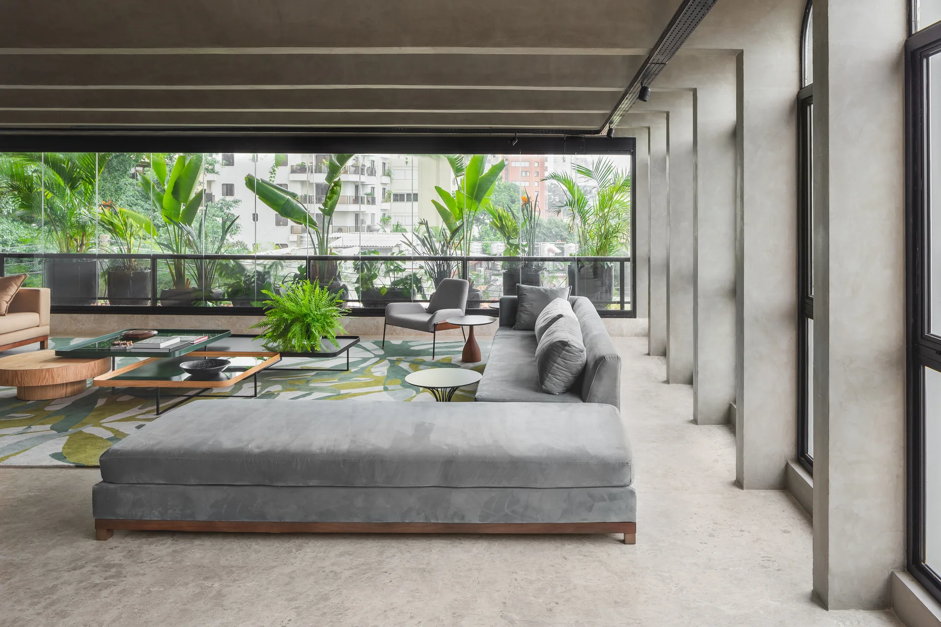 A modern living room features a large gray sectional sofa, a botanical green rug, and concrete columns next to a glass wall showing lush outdoor plants.