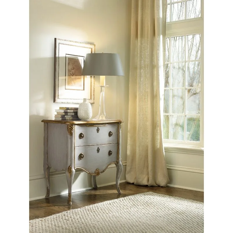 A distressed grey French Provincial chest with gold trim sits near a window with beige linen drapes, topped with a glass lamp and books.