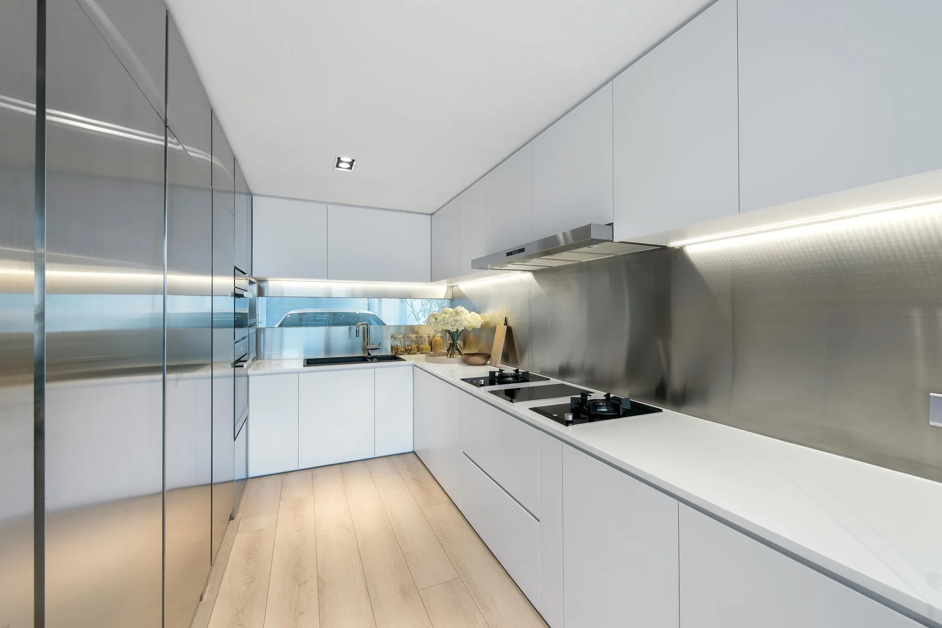 A minimalist galley kitchen features seamless white flat-front cabinets, a brushed stainless steel backsplash, and light wood plank flooring.