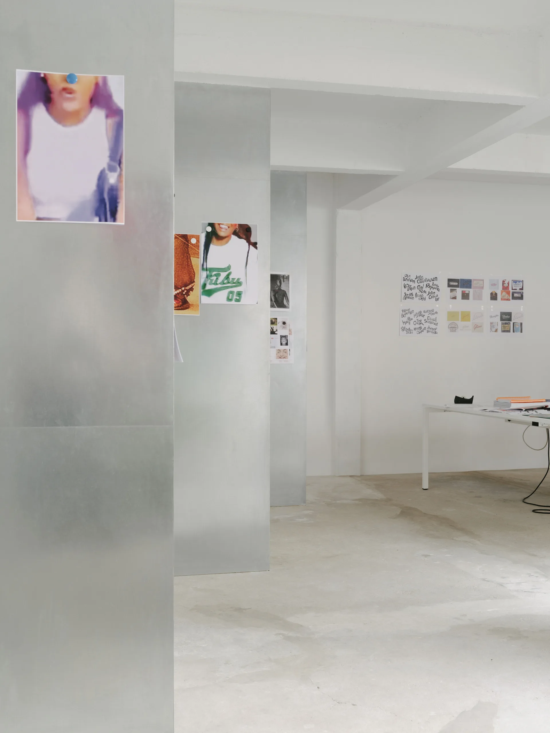 A minimalist industrial studio features large silver metal partitions used as pinboards alongside white walls, concrete flooring, and a white work table.