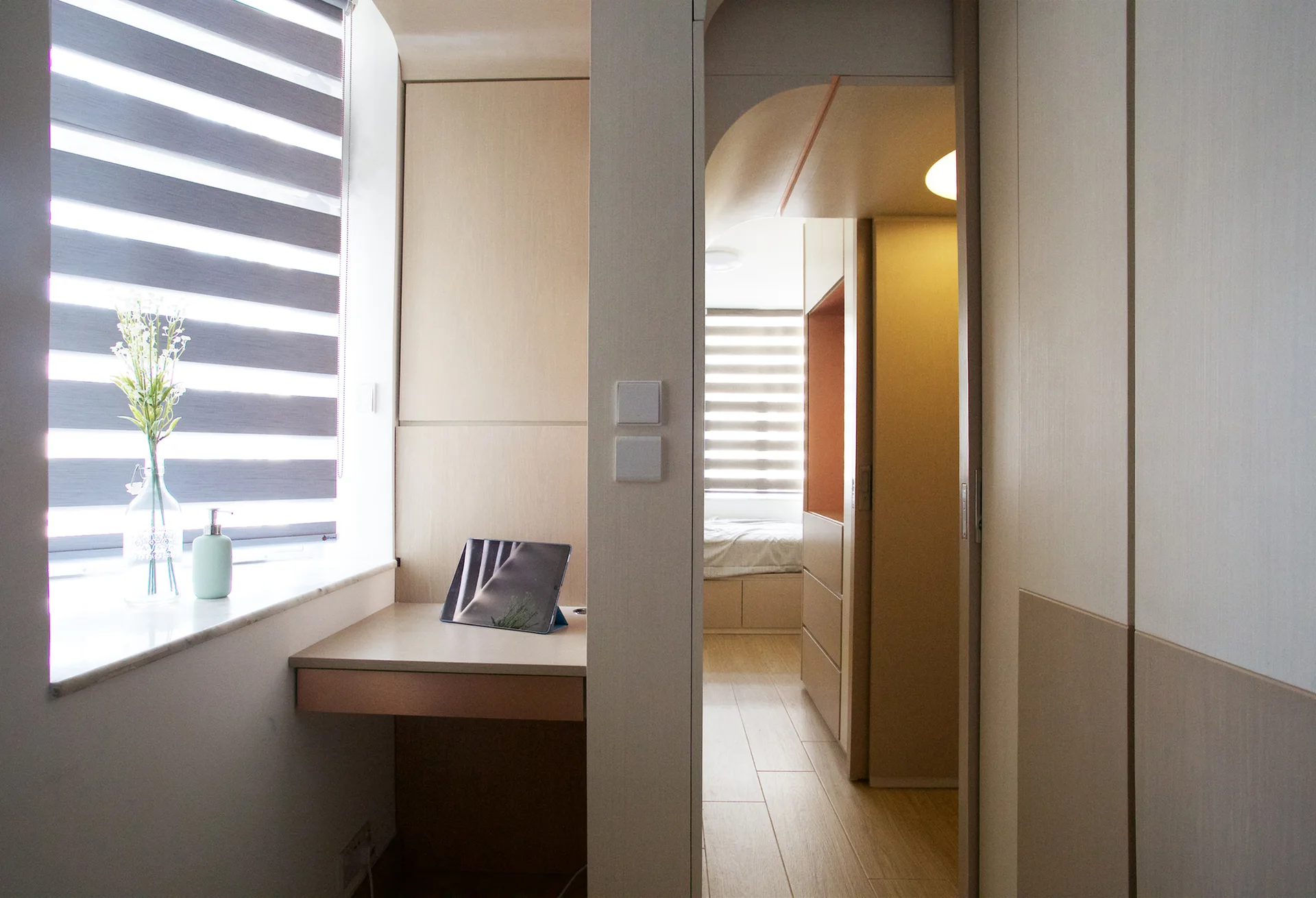 A built-in wooden desk sits below a window with striped blinds next to an open doorway leading to a bedroom with integrated wardrobes.