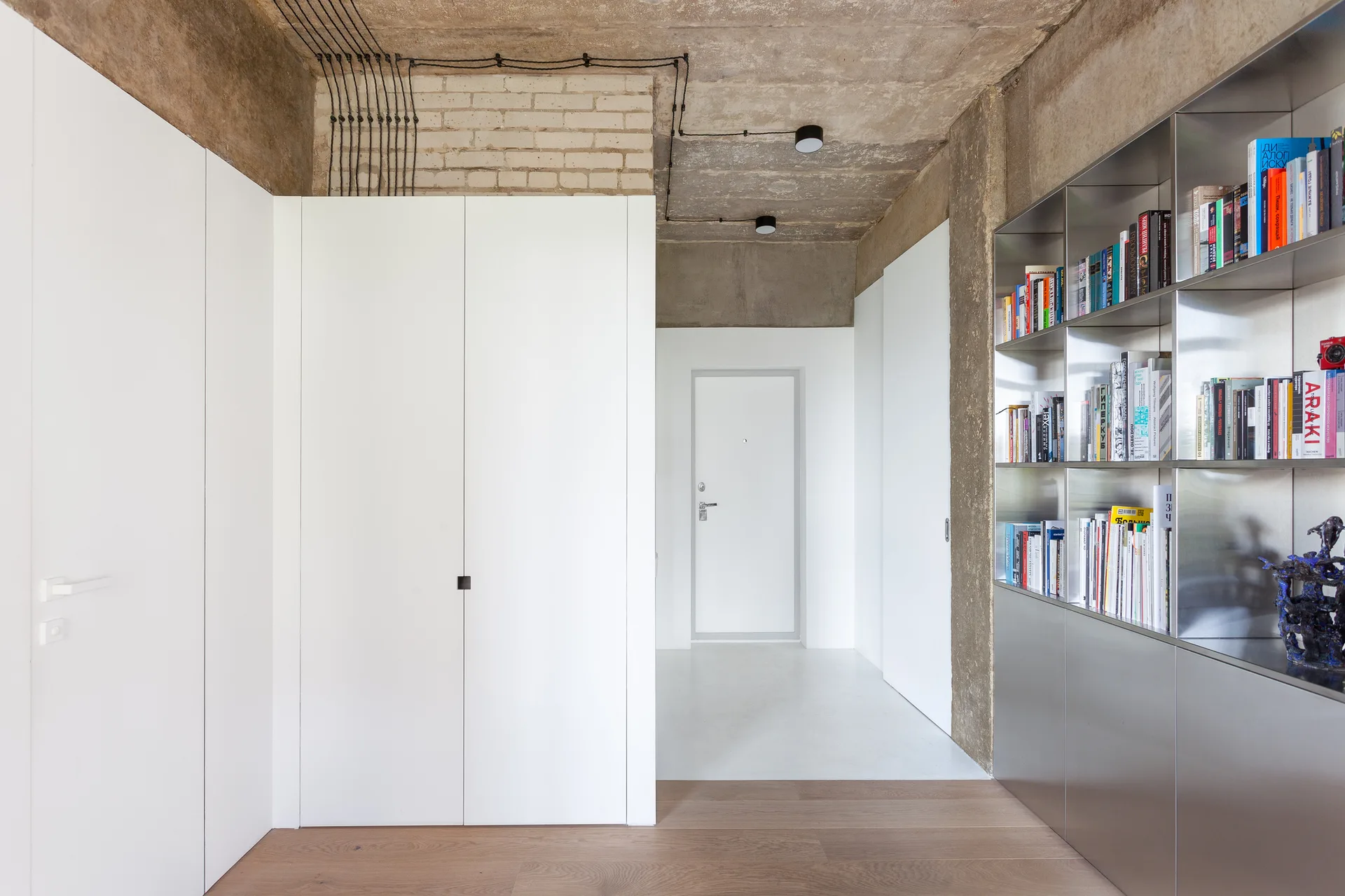 A modern residential entryway featuring minimalist white closets, an exposed concrete ceiling with black conduits, and an inset metal bookshelf.