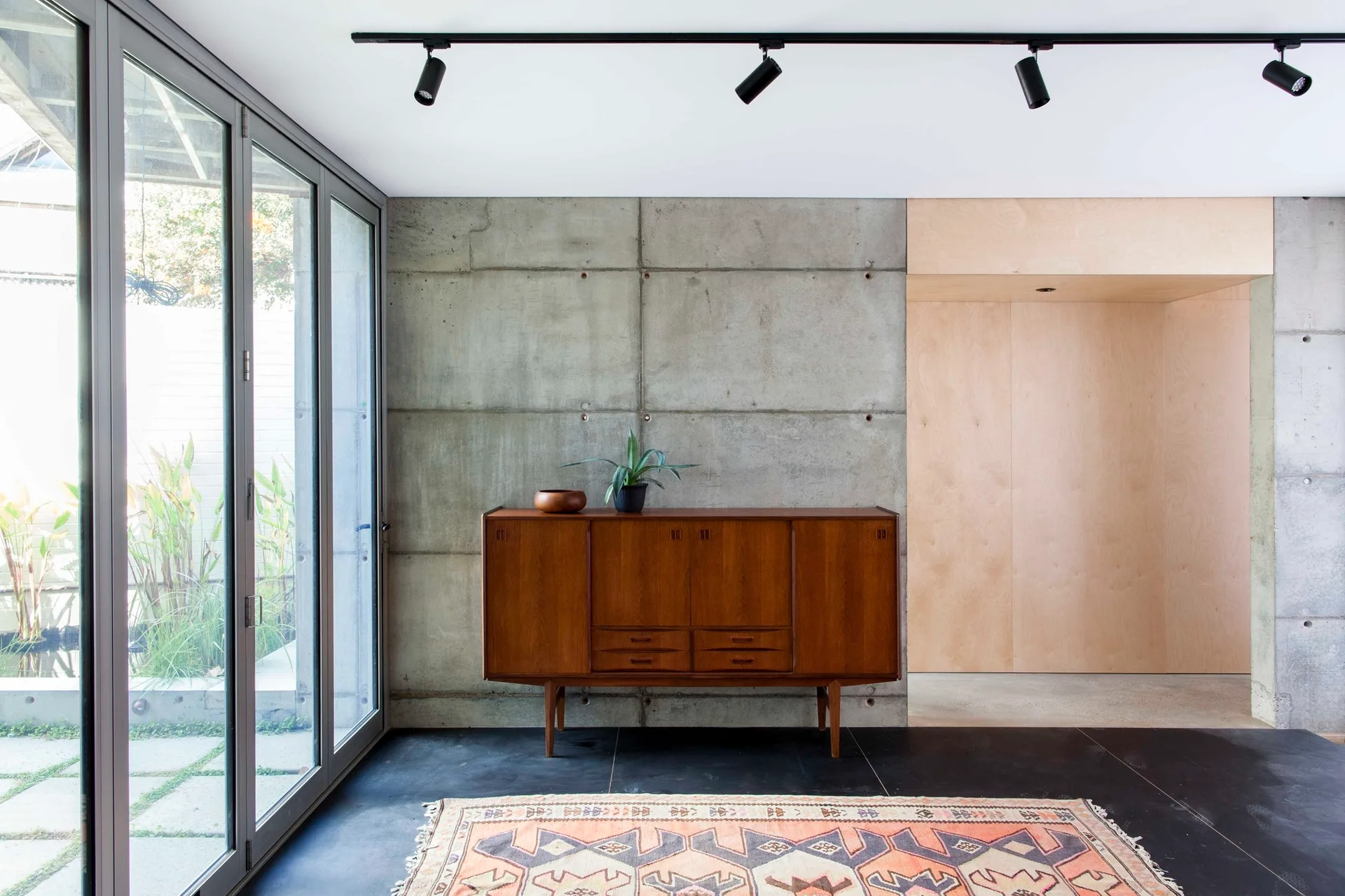A wooden mid-century sideboard stands against a concrete paneled wall next to glass sliding doors and dark tiled flooring.