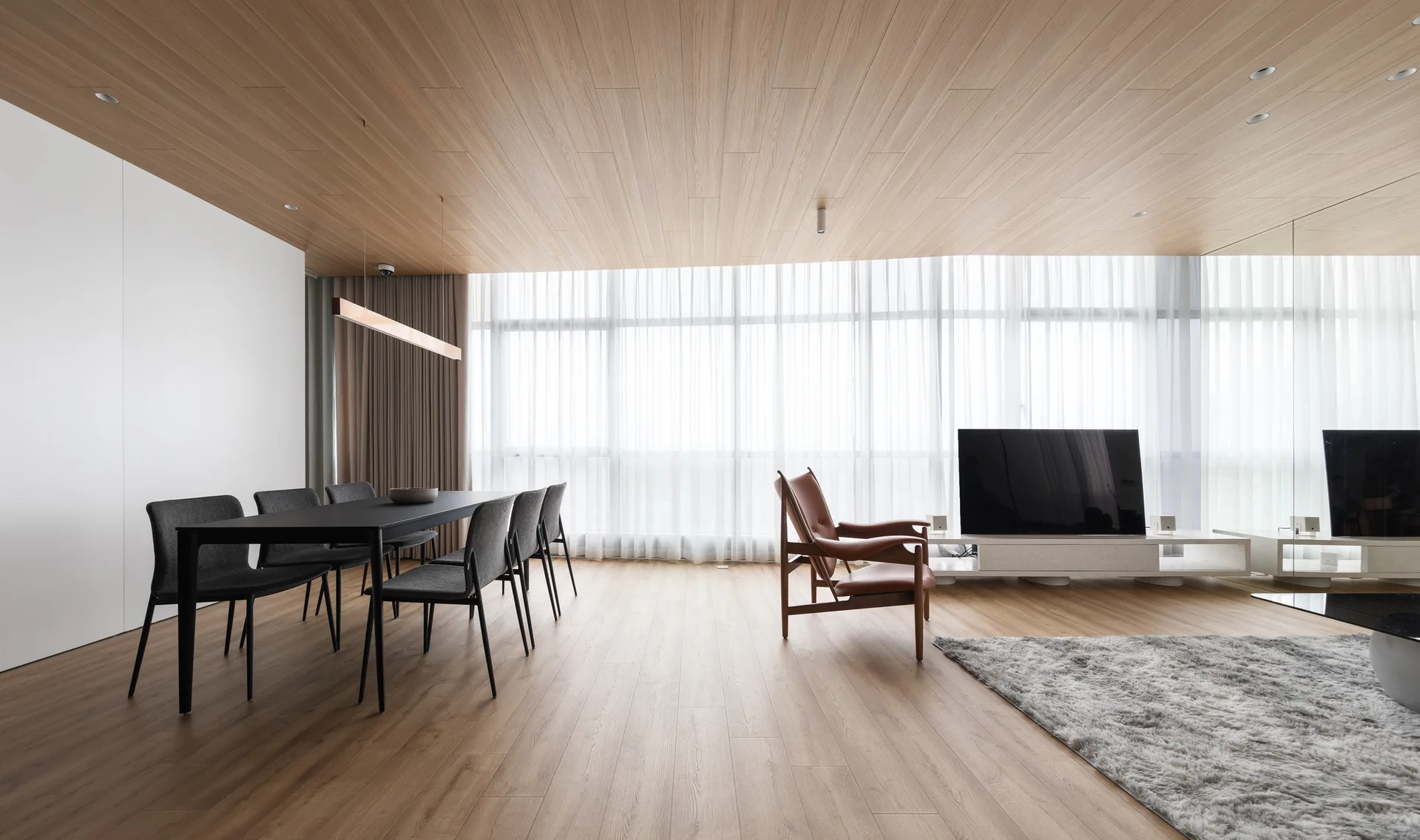 An open-concept living and dining room with a wood ceiling and floor, a black dining set, a brown leather armchair, and sheer white curtains.