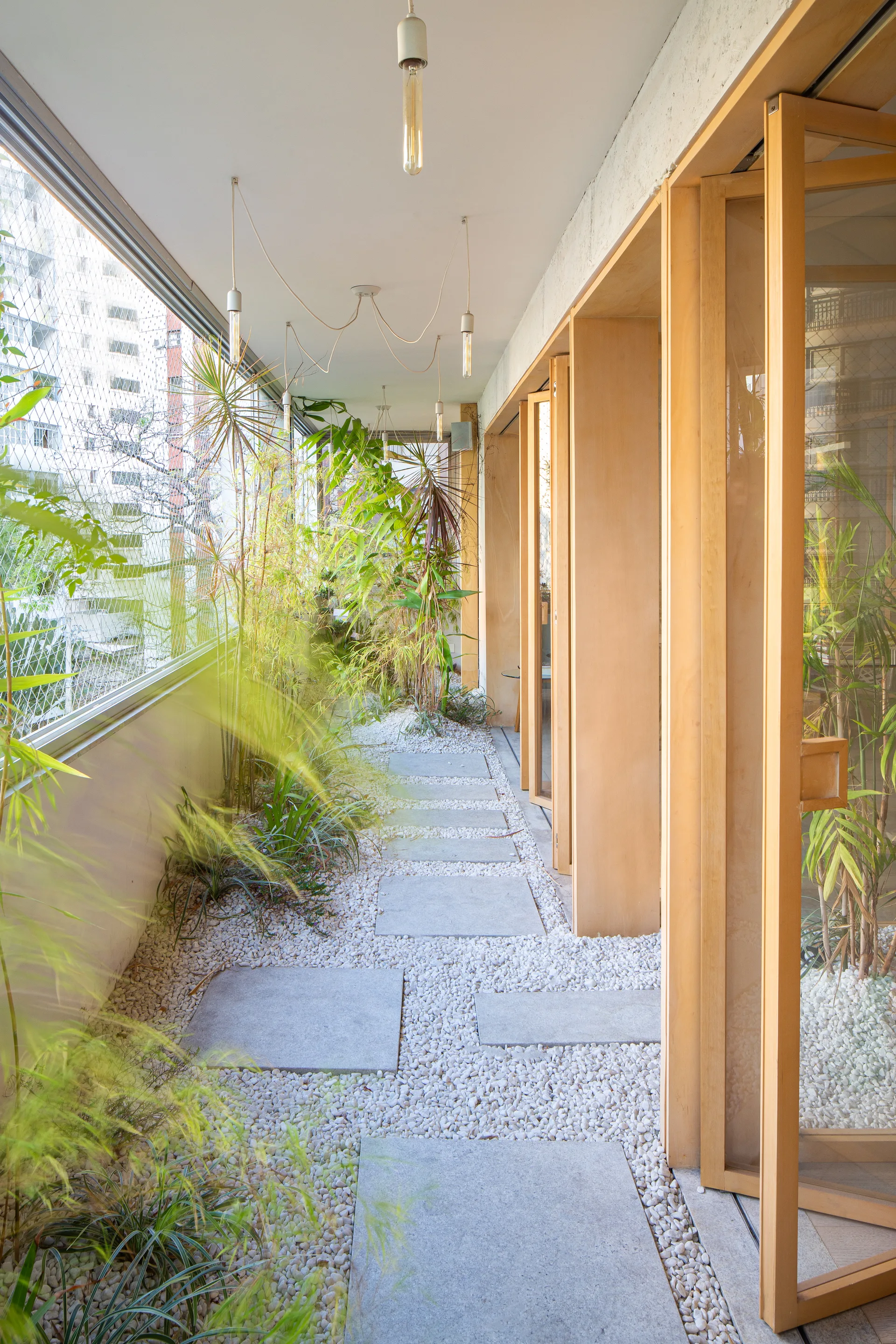 A covered balcony features grey concrete stepping stones set in white gravel alongside lush green plants and light wood door frames.