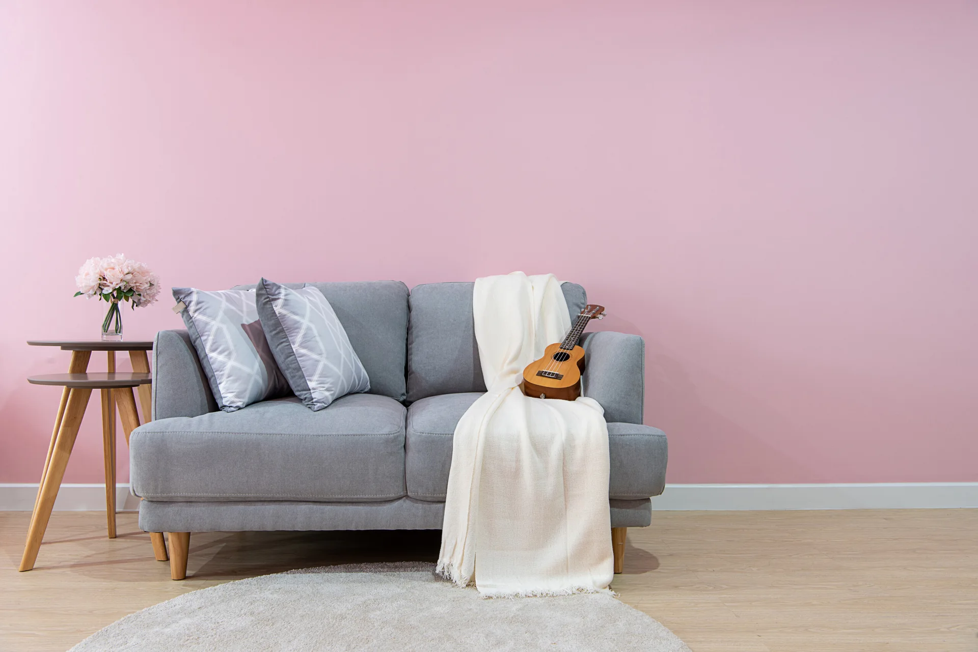 A grey upholstered sofa with patterned pillows sits against a solid pink wall next to a wooden side table with pink flowers.