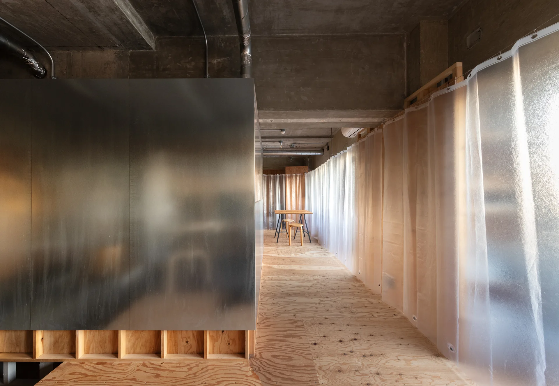 An industrial loft interior featuring a large brushed steel partition wall, a raw concrete ceiling, unfinished plywood floors, and translucent curtains.