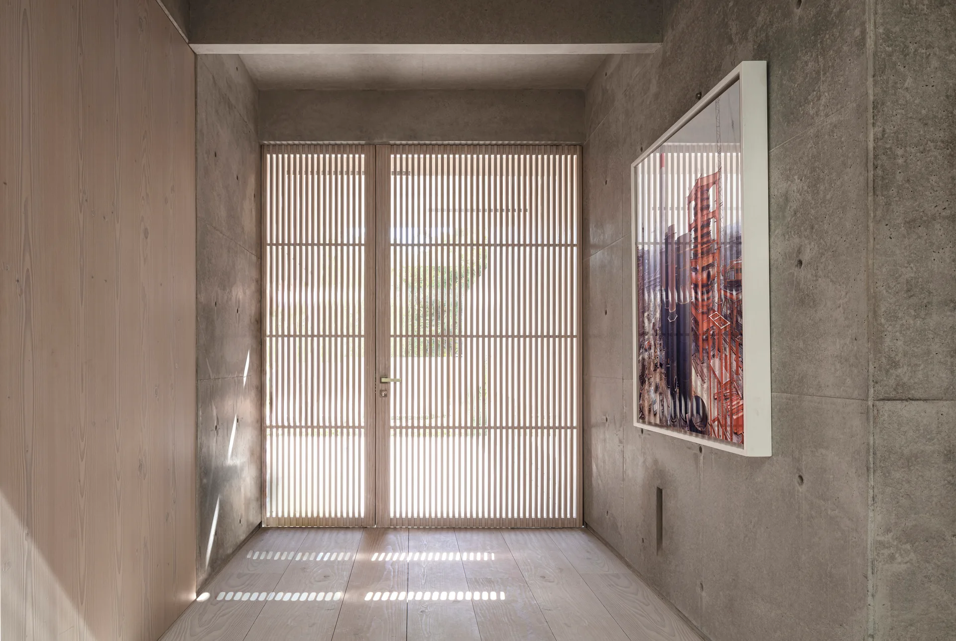 An entryway with exposed concrete walls, a vertical light wood panel, blonde wood flooring, and double slatted wooden doors filtering natural light.