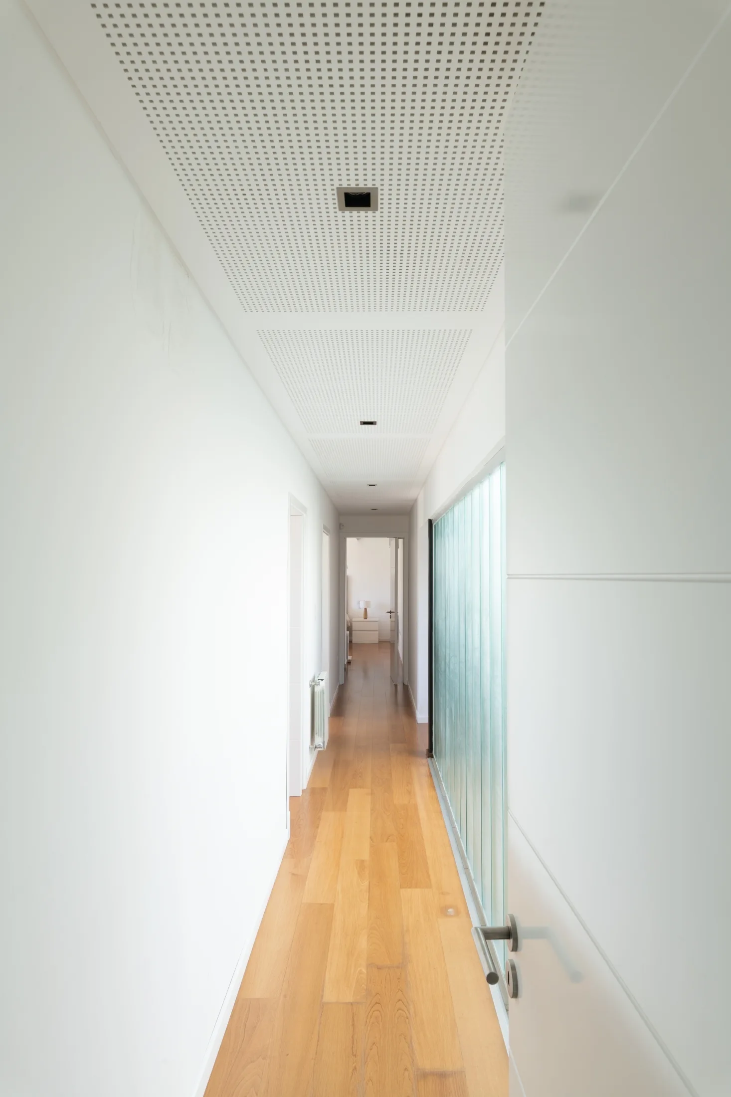 A long minimalist white hallway with light hardwood floors, a perforated grid pattern ceiling, and a textured glass partition.