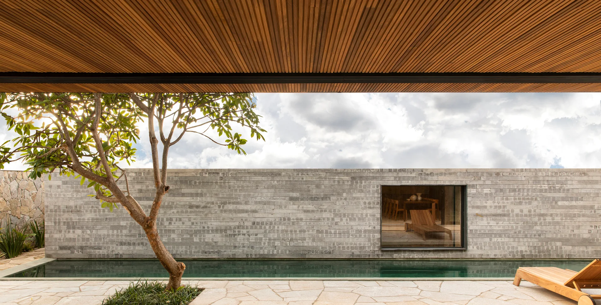 An outdoor pool patio features a long lap pool, a wooden lounge chair, a timber slatted canopy, and a stone wall with a window viewing the interior.