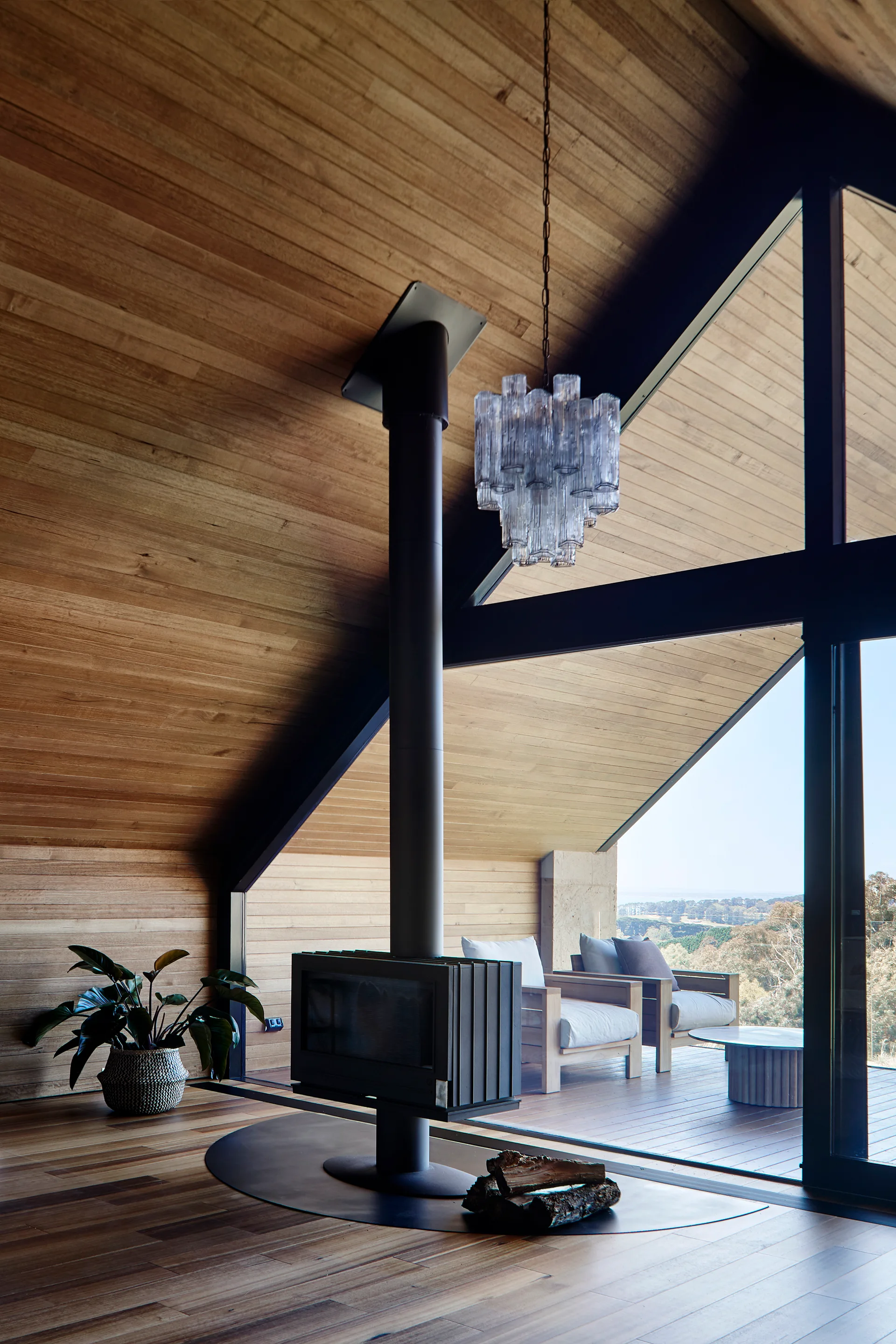 A modern living room with slanted wood ceilings features a black freestanding fireplace, a tiered glass pendant, and large windows opening to a deck.