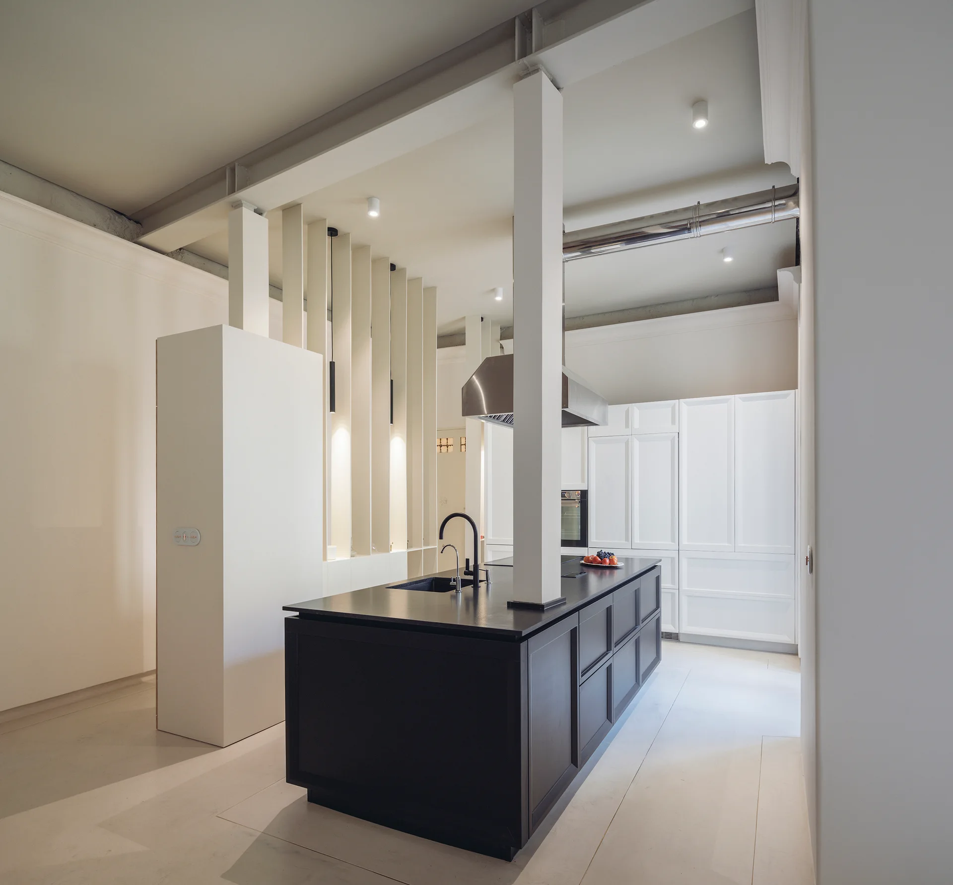 A modern minimalist kitchen featuring a dark central island integrated with a white structural pillar and surrounded by white cabinetry.