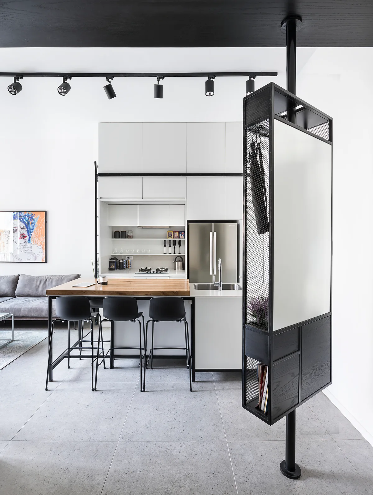 A modern kitchen features a wood-top island with black bar stools, white cabinets, and a tall black metal partition screen.