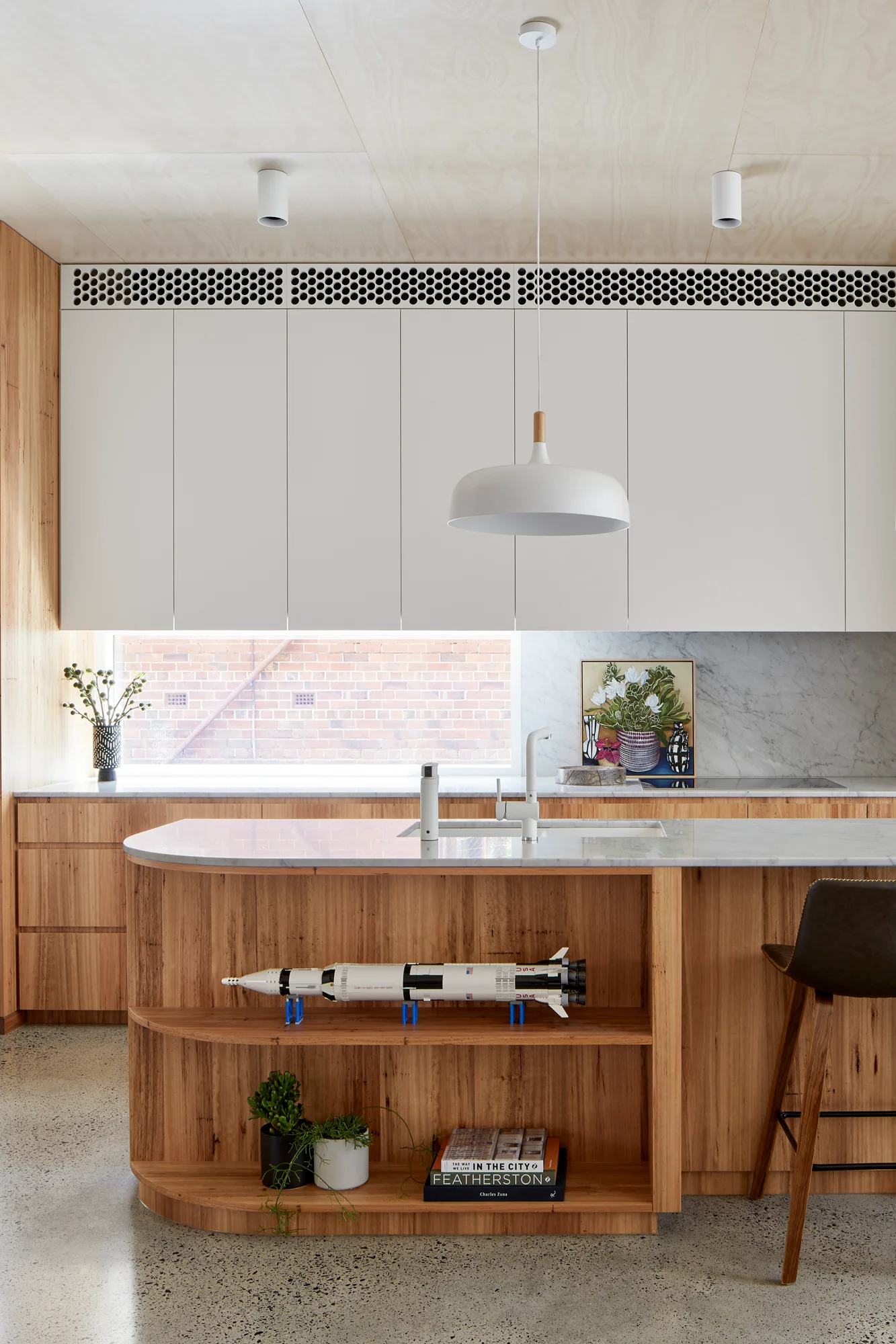 A modern kitchen featuring a natural wood island with curved open shelves, marble countertops, white upper cabinets, and a horizontal backsplash window.
