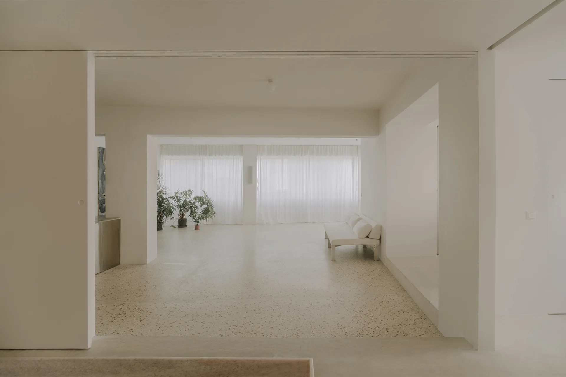 A minimalist open-plan room with light beige terrazzo flooring, a white low-profile sofa, and sheer white curtains covering a large window.