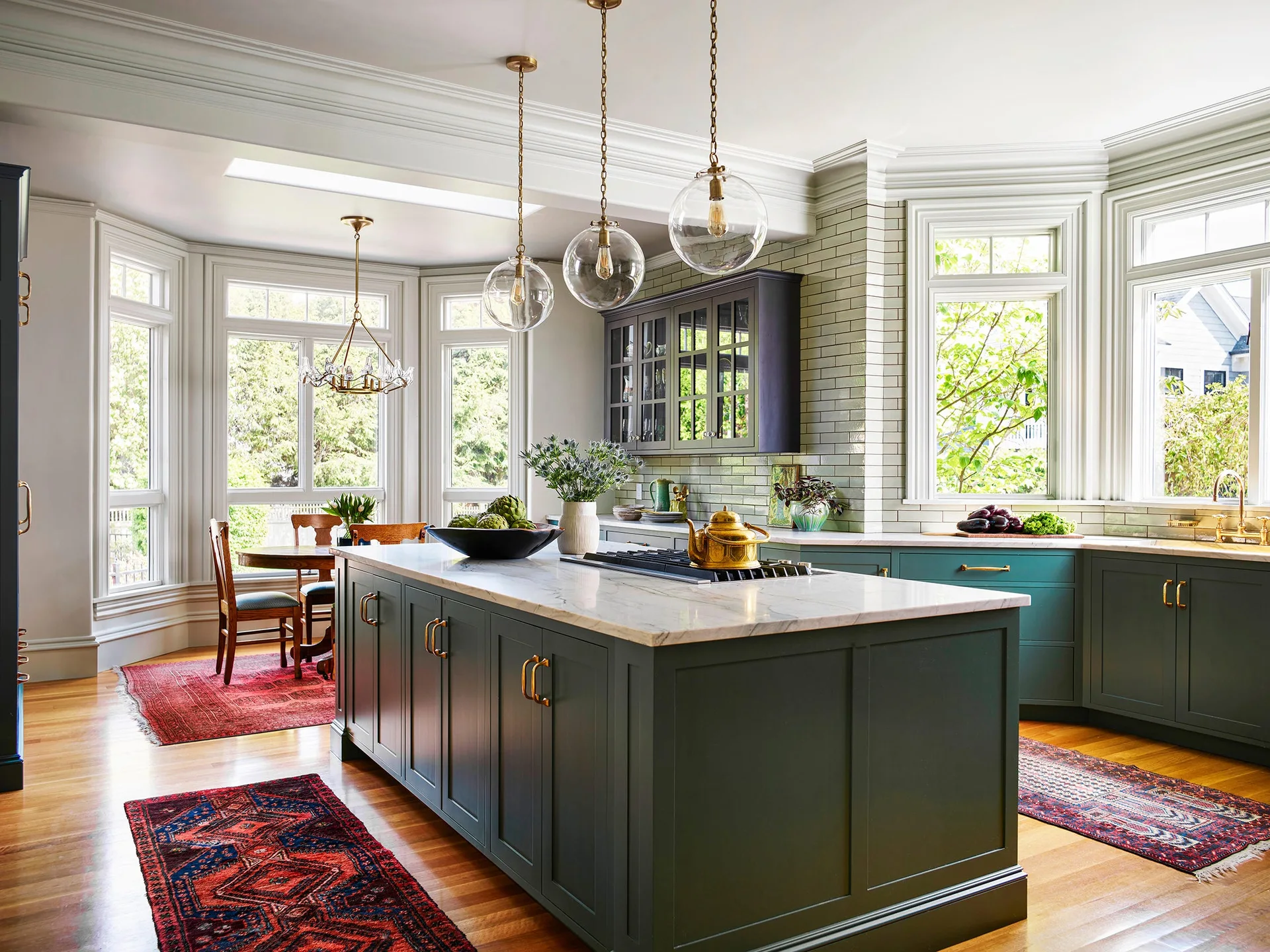 A spacious kitchen features dark green cabinets, a marble-topped center island, three hanging glass globe lights, and vintage runner rugs on wood floors.