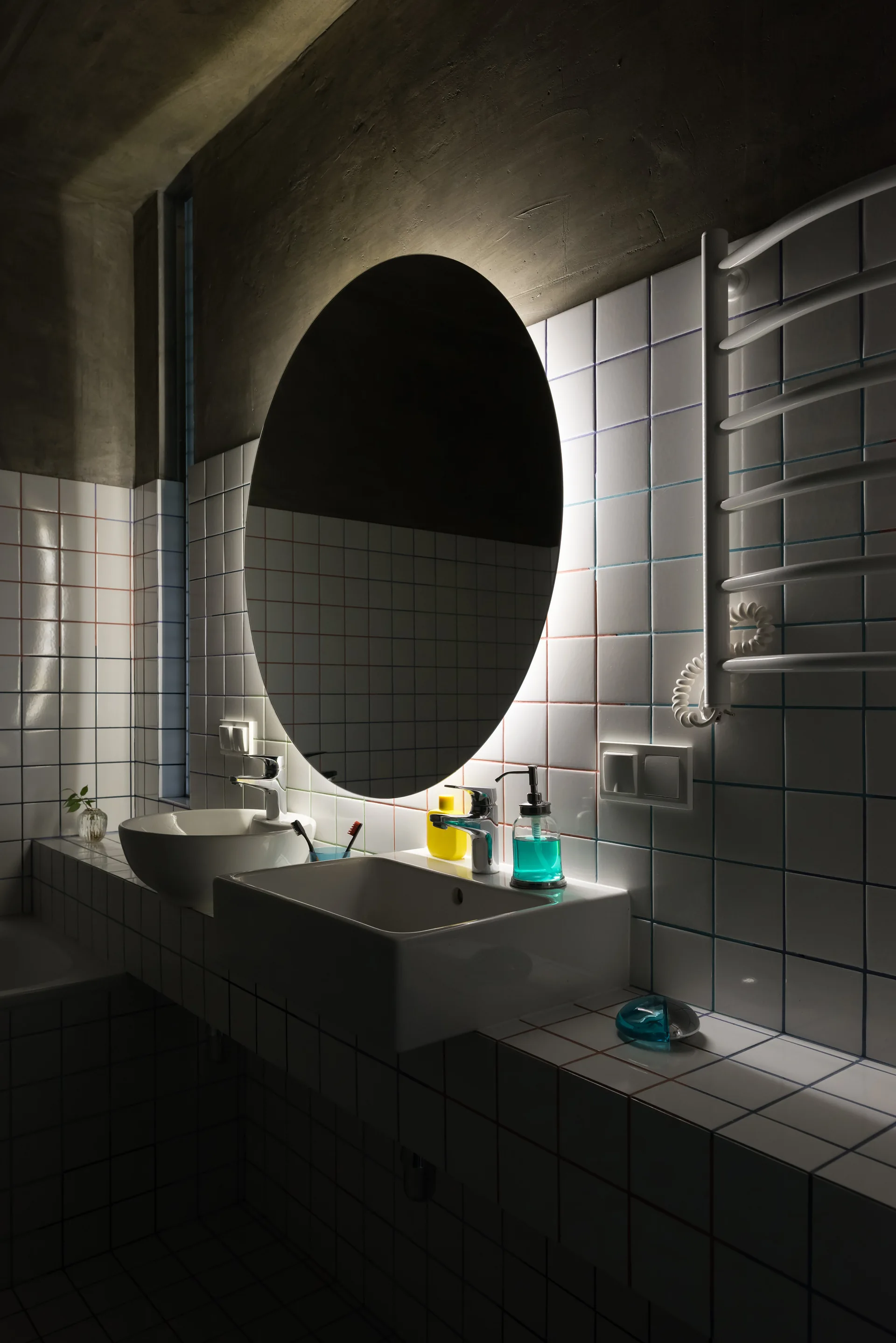 A modern bathroom features a tiled vanity with two white sinks, a large backlit round mirror, and a raw concrete ceiling.