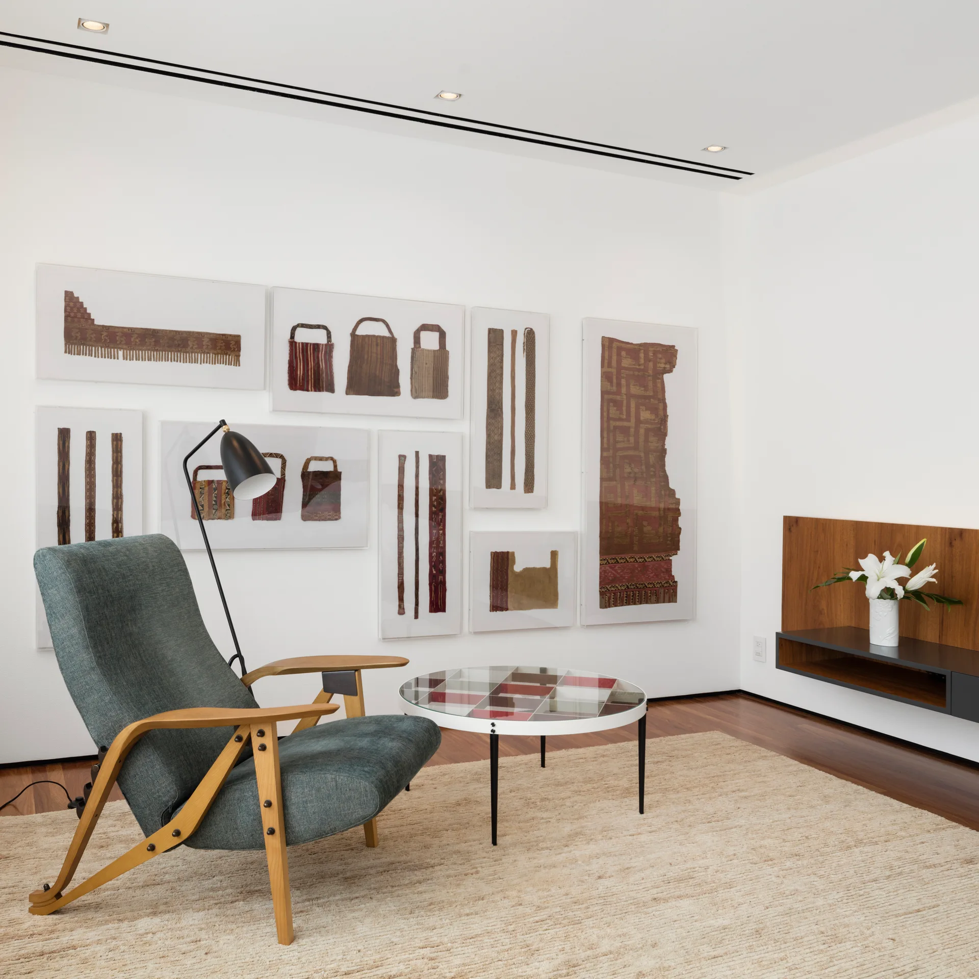 A blue-grey mid-century armchair and patterned coffee table sit on a woven rug in front of a white wall decorated with framed textile art.