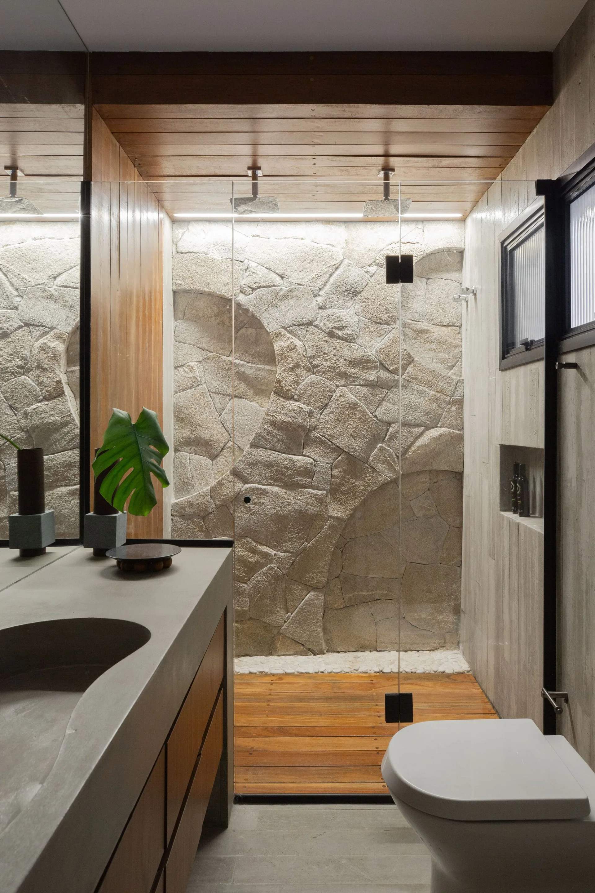 A modern organic bathroom showcases a glass-enclosed shower with a textured stone wall, wood ceiling and floor planks, and a concrete vanity.